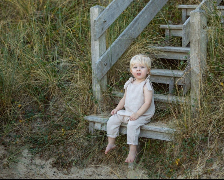 blonde toddler boy sits on the wooden steps on the beach wearing his bohemian hippy style adjustable romper in the colour Natural sustainable baby clothing