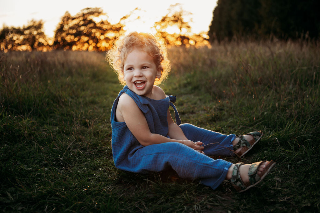 toddler boy sat in a meadow sunset behind wearing handmade bohemian romper with adjustable straps in blue curly hair hippy style clothing unisex eco friendly sustainable