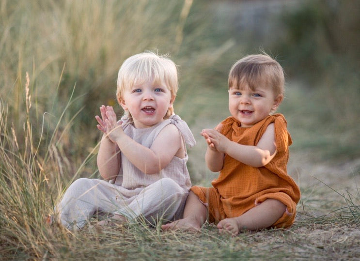 two brothers sat together on the sand wearing handmade rompers in orange and natural clapping their hands toddler and baby bohemian hippy style unisex