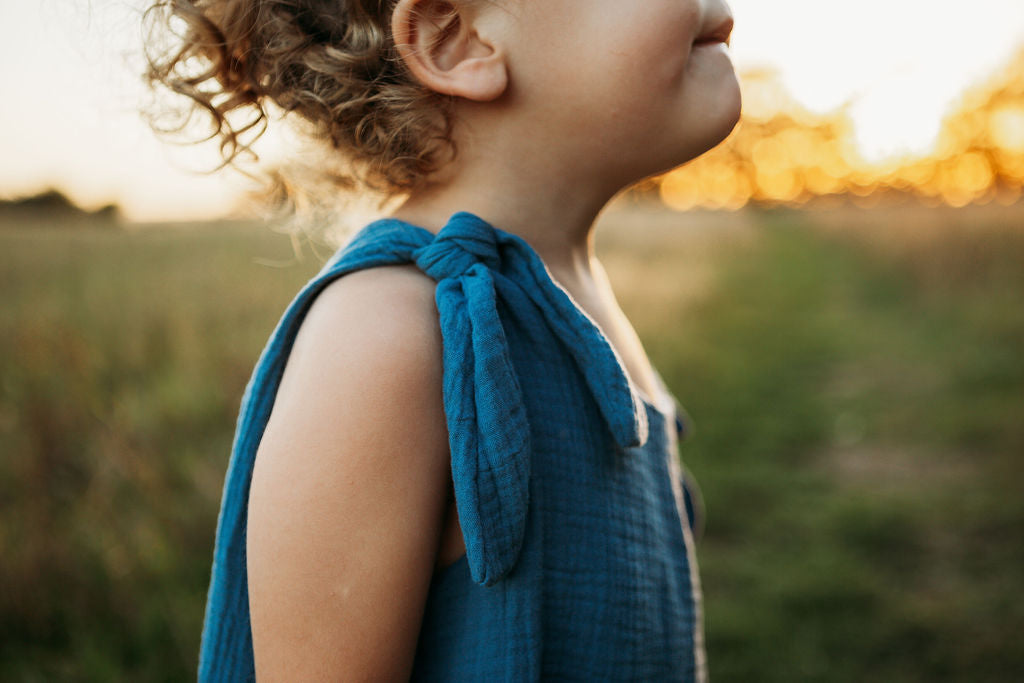 toddler boy stood in a meadow sunset behind wearing handmade bohemian romper with adjustable straps in blue curly hair hippy style clothing unisex eco friendly sustainable 