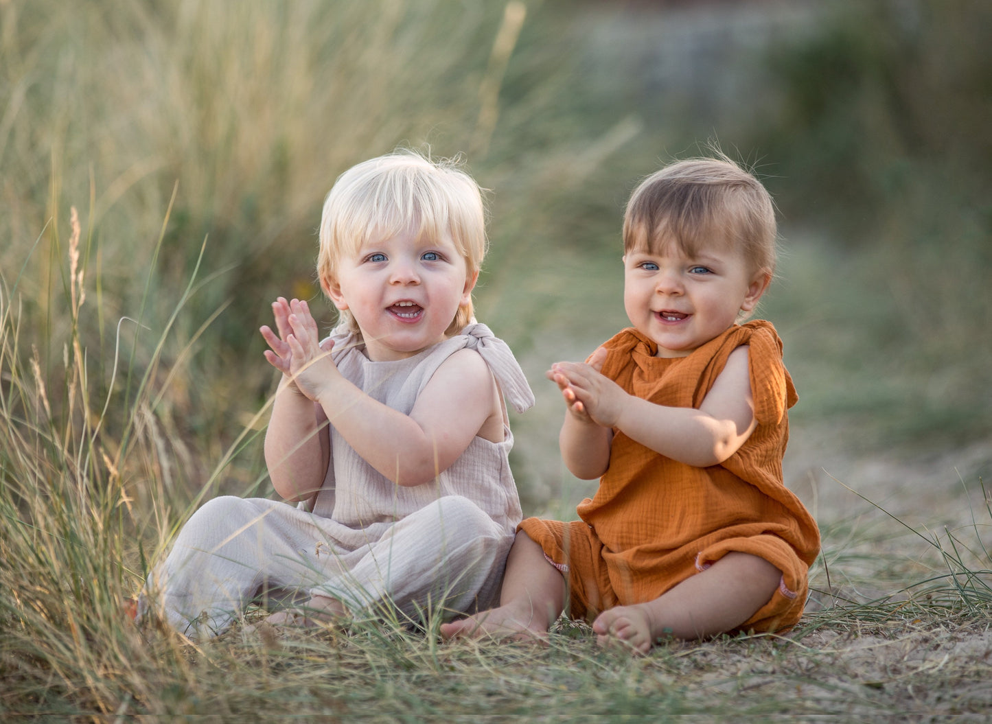 two brothers sat together on the sand wearing handmade rompers in orange and natural clapping their hands toddler and baby bohemian hippy style unisex  