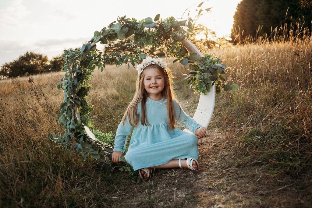 little girl sat in flower arch in a bohemain meadow with sunset in the background wearing a flower crown wearing her glitter sparkle handmade jersey dress party disco occasion dress inspired frozen elsa sustainable