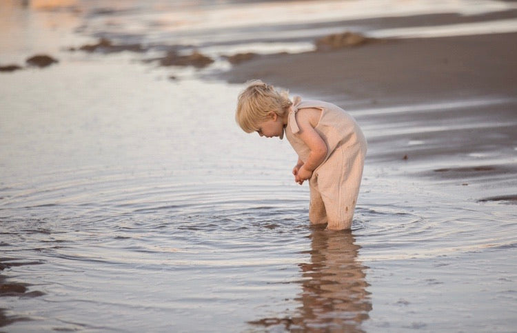 blonde toddler boy walks along the beach bending into the sea splashing wearing his bohemian hippy style adjustable romper in the colour Natural sustainable baby clothing