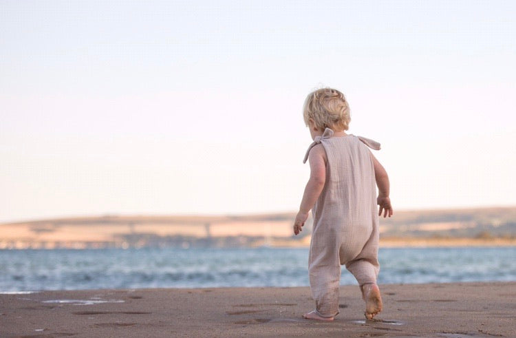 blonde toddler boy walks along the beach with the sea in the background wearing his bohemian hippy style adjustable romper in the colour Natural sustainable baby clothing